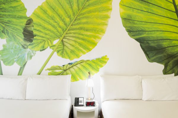 The image shows two beds with white bedding, separated by a small table with a lamp, phone, and alarm clock. Large leaf artwork is on the wall.