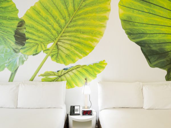 The image shows two beds with white bedding, separated by a small table with a lamp, phone, and alarm clock. Large leaf artwork is on the wall.