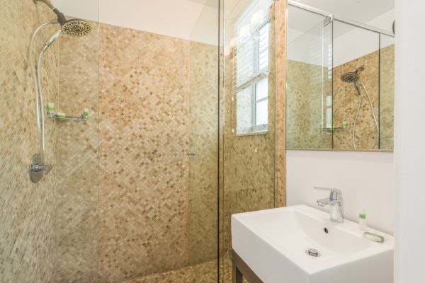 The image shows a bathroom with a glass-enclosed shower, a mosaic tile wall, a sink with a faucet, a mirror, and a small bottle on the sink counter.