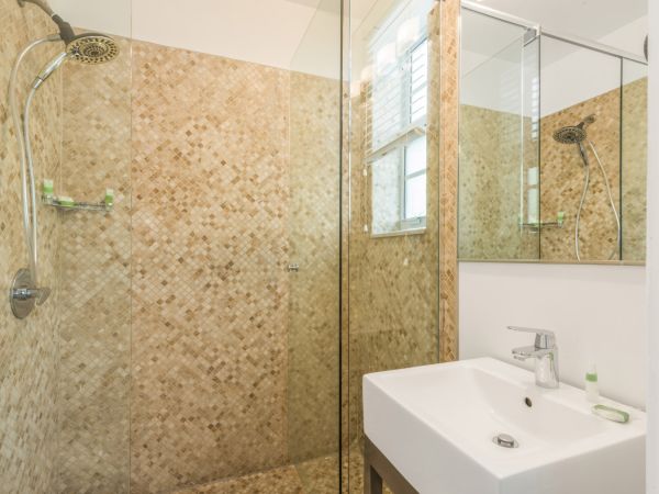 The image shows a bathroom with a glass-enclosed shower, a mosaic tile wall, a sink with a faucet, a mirror, and a small bottle on the sink counter.