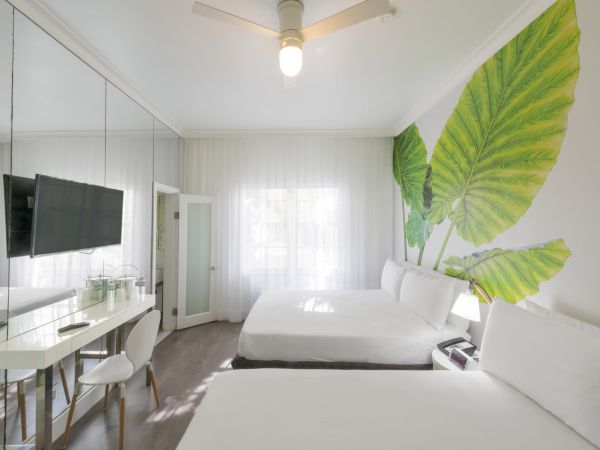 The image shows a modern hotel room with two beds, a wall-mounted TV, a desk, chairs, and leaf-themed wall decor, ending the sentence.