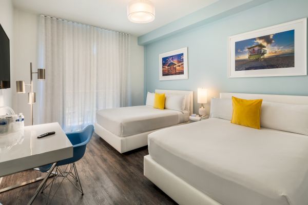 The image shows a neatly arranged hotel room with two double beds, a desk with a chair, a TV, and framed artwork on pale blue walls.