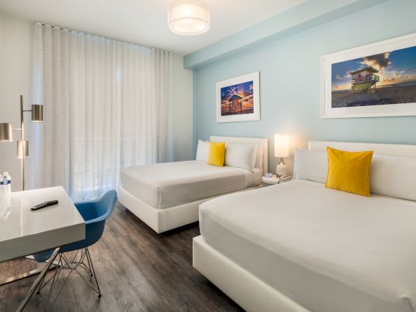The image shows a neatly arranged hotel room with two double beds, a desk with a chair, a TV, and framed artwork on pale blue walls.