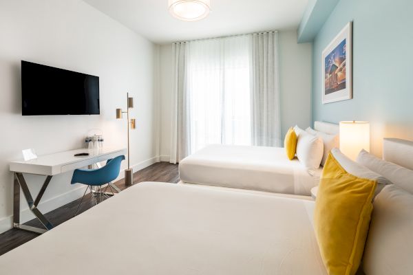 A modern hotel room with two beds, yellow pillows, a wall-mounted TV, a desk with a chair, and large windows with sheer curtains.