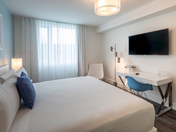 A modern bedroom with a large bed, a desk with a blue chair, a wall-mounted TV, and a window with white curtains.