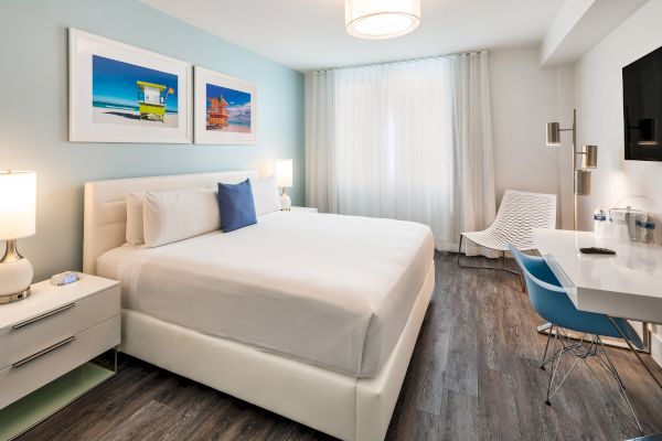 This image shows a modern bedroom with a large bed, white linens, two bedside tables, a desk, and a chair. Decor includes two colorful photos.