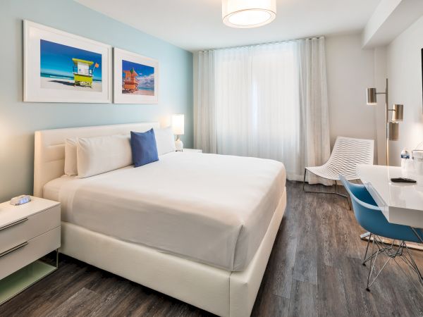 This image shows a modern bedroom with a large bed, white linens, two bedside tables, a desk, and a chair. Decor includes two colorful photos.