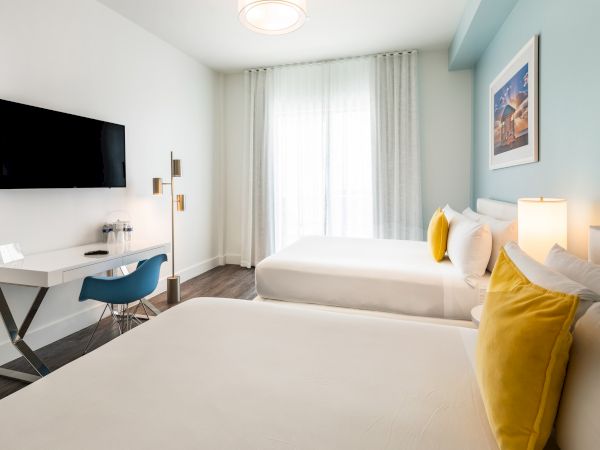 A modern hotel room featuring two beds with white linens and yellow pillows, a wall-mounted TV, a work desk with a chair, and a window with sheer curtains.
