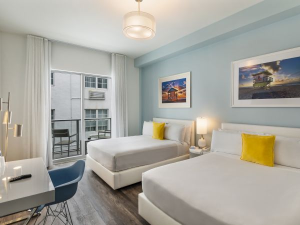 A hotel room with two double beds, yellow pillows, wall art, and a desk beside a window with a view of neighboring buildings.