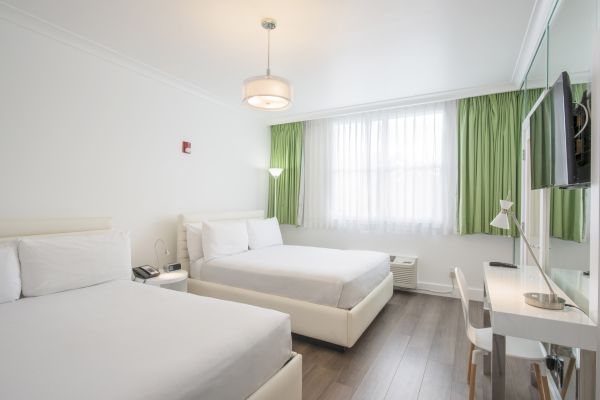 The image shows a clean, modern hotel room with two double beds, white linens, green curtains, a desk, a wall-mounted TV, and a hardwood floor.