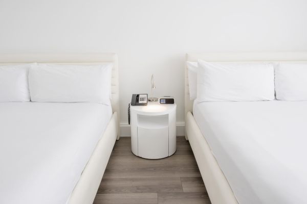 The image shows two white beds with a nightstand between them. On the nightstand are a lamp, a telephone, and a digital clock.