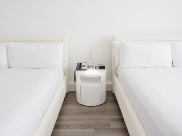 The image shows two white beds with a nightstand between them. On the nightstand are a lamp, a telephone, and a digital clock.