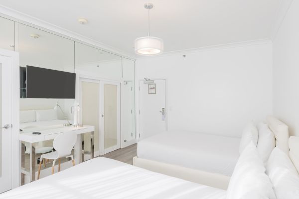 The image shows a bright, minimalist hotel room with two beds, a desk and chair, a wall-mounted TV, and a full-length mirror on the wall.