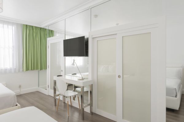 A modern bedroom with green curtains, a wall-mounted TV, a small desk with a chair, and white sliding doors.