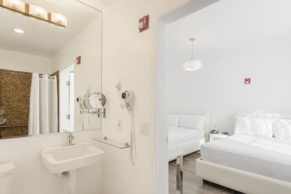 A bathroom with a sink, mirror, hairdryer, and a view into a bedroom with a bed, nightstand, lamp, and minimal decor, all in white.