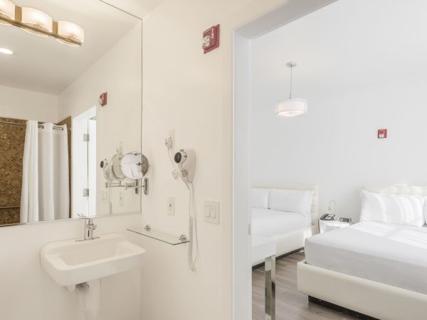 A bathroom with a sink, mirror, hairdryer, and a view into a bedroom with a bed, nightstand, lamp, and minimal decor, all in white.