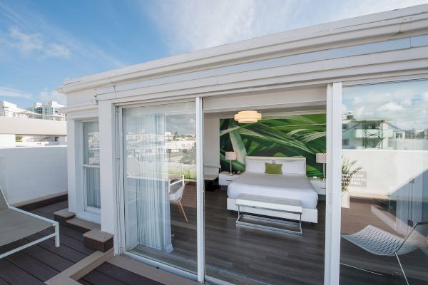 A modern room with large glass doors opens to a deck with lounge chairs. Inside, there's a bed with a green-themed wall art and a hanging light.