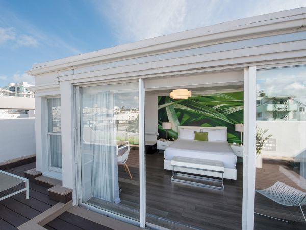 A modern room with large glass doors opens to a deck with lounge chairs. Inside, there's a bed with a green-themed wall art and a hanging light.