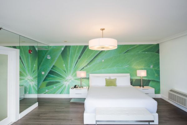 A modern bedroom with a green leaf mural, white bed, bedside tables, lamps, and a mirrored wall on the left.