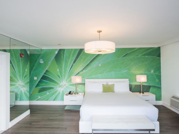 A modern bedroom with a green leaf mural, white bed, bedside tables, lamps, and a mirrored wall on the left.