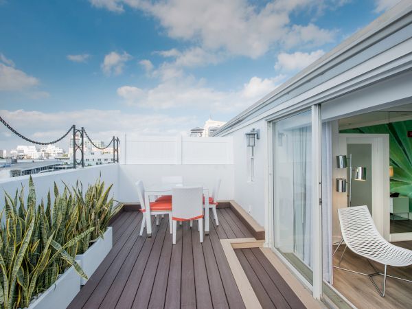 A rooftop terrace with outdoor seating, potted plants, and a sliding door leading to an interior space with modern decor under a partly cloudy sky.