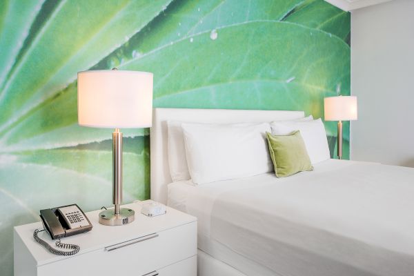 A modern bedroom features a white bed, two lamps, a green leaf wall mural, a nightstand with a phone, and a small green pillow.