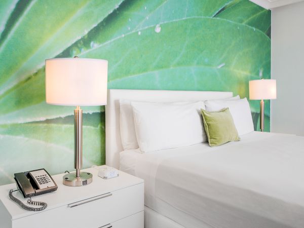 A modern bedroom features a white bed, two lamps, a green leaf wall mural, a nightstand with a phone, and a small green pillow.