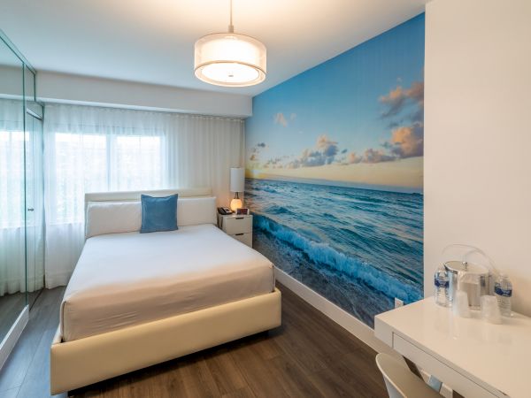 A modern bedroom with a bed, mirrored wall, ocean-themed wall mural, desk, chair, and pendant light.