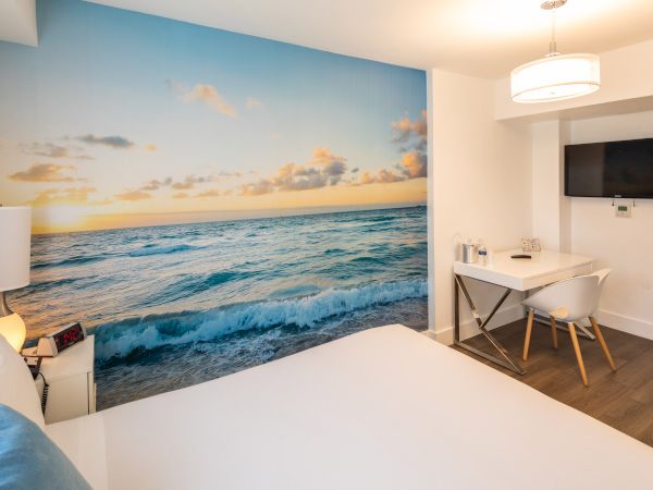 A modern hotel room with a large beach mural, a bed, desk with a chair, lamp, and a mounted flat-screen TV on the wall always ending the sentence.