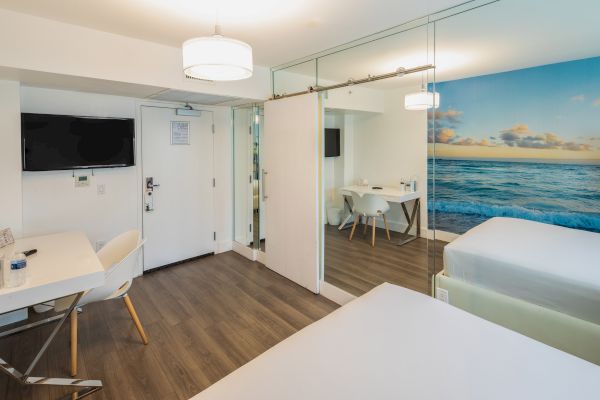 A modern hotel room featuring two beds, a desk, a TV, and a partitioned seating area with a table. A beach mural adds color to the space.