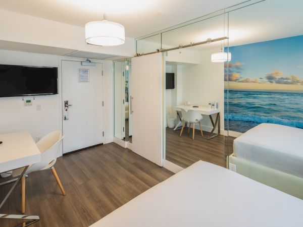 A modern hotel room featuring two beds, a desk, a TV, and a partitioned seating area with a table. A beach mural adds color to the space.