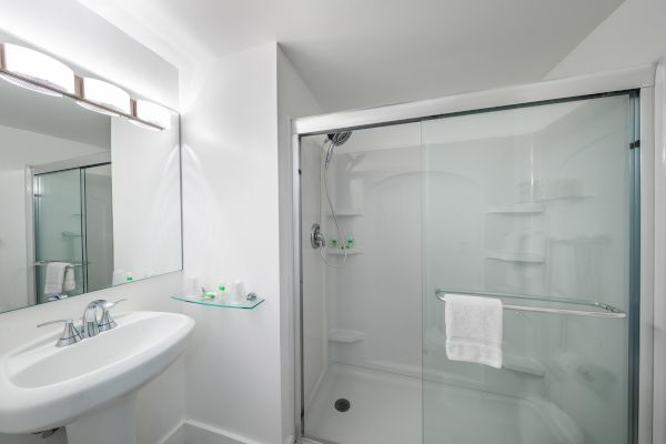 A modern bathroom with a glass-enclosed shower, white sink, and a wall-mounted mirror above the sink. Shelves in the shower hold toiletries.