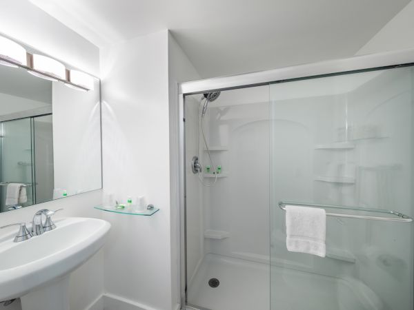 A modern bathroom with a glass-enclosed shower, white sink, and a wall-mounted mirror above the sink. Shelves in the shower hold toiletries.