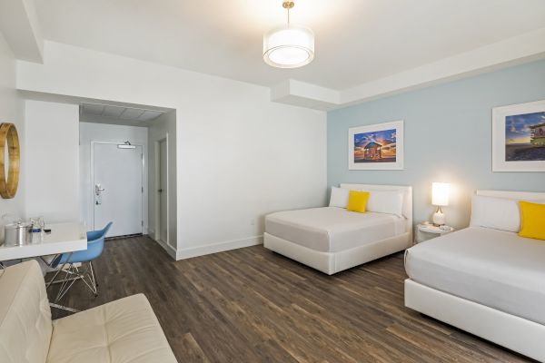 A modern hotel room with two beds, two wall paintings, a small desk, a chair, a sofa, and wooden flooring.