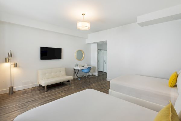 A modern hotel room with two beds, a wall-mounted TV, a small sofa, a desk with a chair, a round mirror, and a tall lamp on wooden flooring.