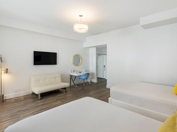A modern hotel room with two beds, a wall-mounted TV, a small sofa, a desk with a chair, a round mirror, and a tall lamp on wooden flooring.