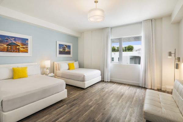 A modern, bright room features two beds with yellow pillows, a sofa, wall art, a window, wood flooring, and a ceiling light fixture, ending the sentence.