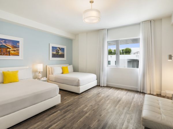 A modern, bright room features two beds with yellow pillows, a sofa, wall art, a window, wood flooring, and a ceiling light fixture, ending the sentence.