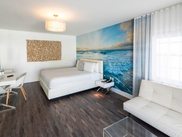 A modern hotel room with a white bed, wall art of an ocean scene, a desk with a chair, a sofa, and a large window with sheer curtains ending the sentence.