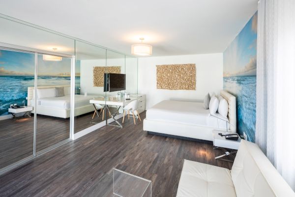 This image shows a modern, clean bedroom with a large mirror, ocean-themed wallpaper, light wooden floors, and white furniture, including a bed and a sofa.