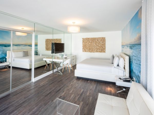 This image shows a modern, clean bedroom with a large mirror, ocean-themed wallpaper, light wooden floors, and white furniture, including a bed and a sofa.
