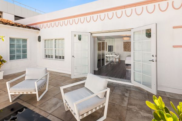 An outdoor patio with two white chairs and a side table leads to an interior room through open double doors in a bright, modern building.