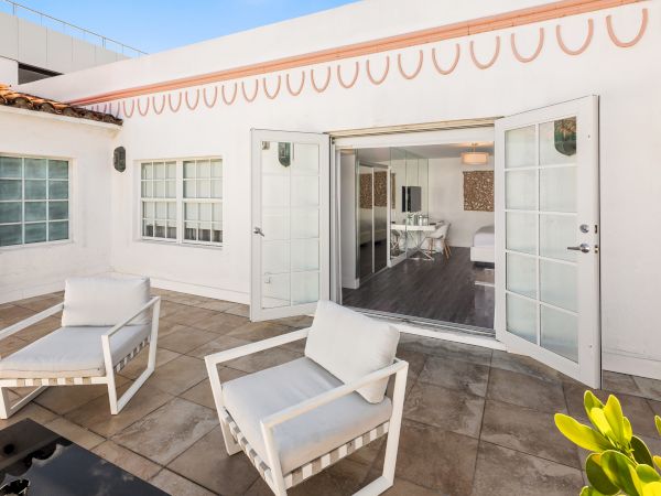An outdoor patio with two white chairs and a side table leads to an interior room through open double doors in a bright, modern building.