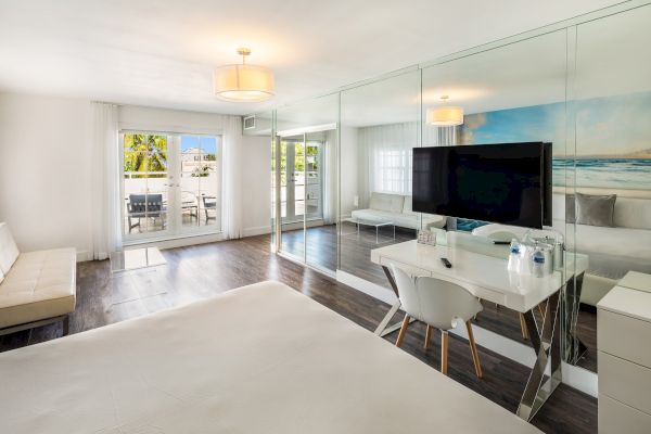 A modern, bright room with a bed, a wall-mounted TV, a desk, mirrored closet doors, and a sliding glass door leading to a patio outside.