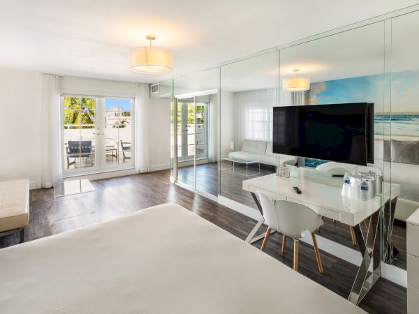 A modern, bright room with a bed, a wall-mounted TV, a desk, mirrored closet doors, and a sliding glass door leading to a patio outside.