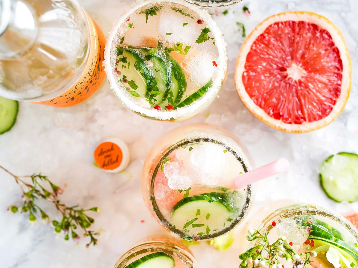 The image depicts a refreshing setup with several drinks garnished with herbs and citrus, a halved grapefruit, cucumber slices, and a bottle.