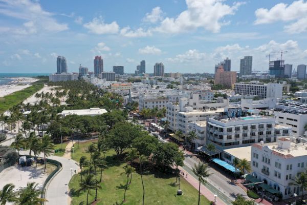 Aerial view of a coastal city with high-rise buildings, parks, and a beach in the distance, featuring palm trees and blue skies ending the sentence.