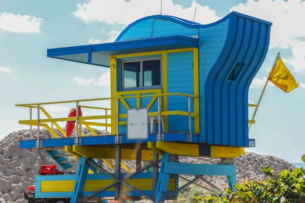 A colorful lifeguard tower painted in blue and yellow with a unique design, a yellow flag, and a life preserver visible against a beach backdrop.