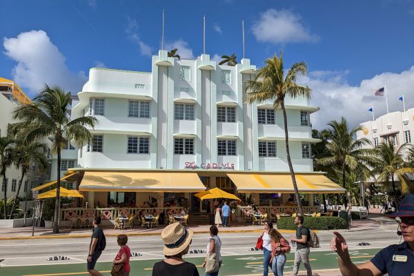 People walking near a white art deco building with yellow awnings, set against a bright blue sky and surrounded by palm trees.