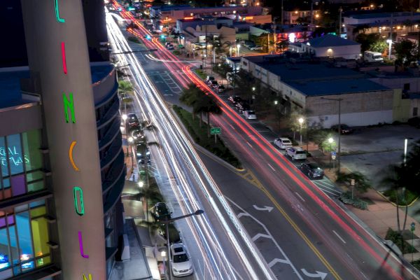 A cityscape at night with streaks of car lights on the road, illuminated buildings, and a colorful vertical sign reading 